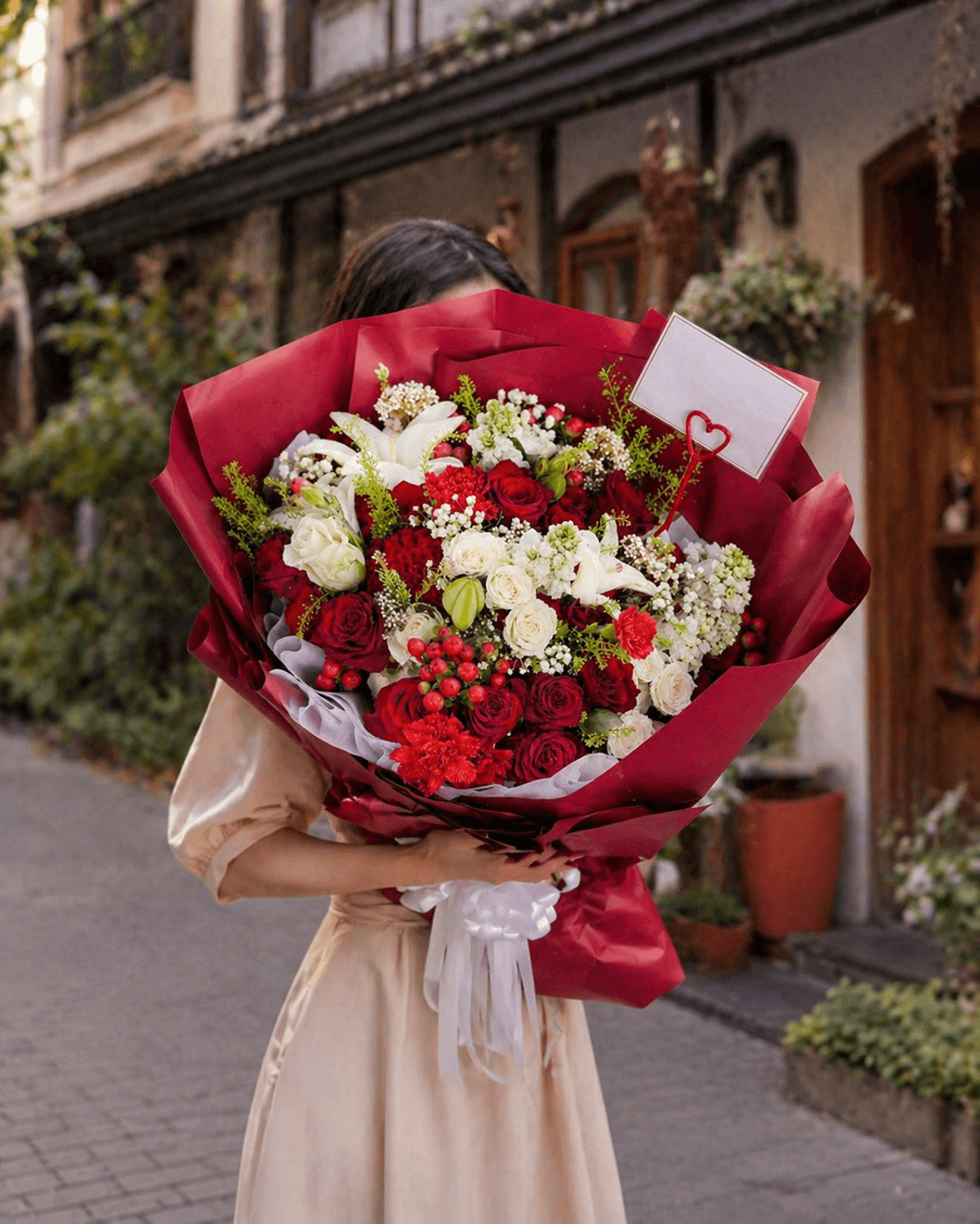 A luxurious romantic bouquet of red roses, white lilies, and red carnations wrapped in dark red paper with a white ribbon.